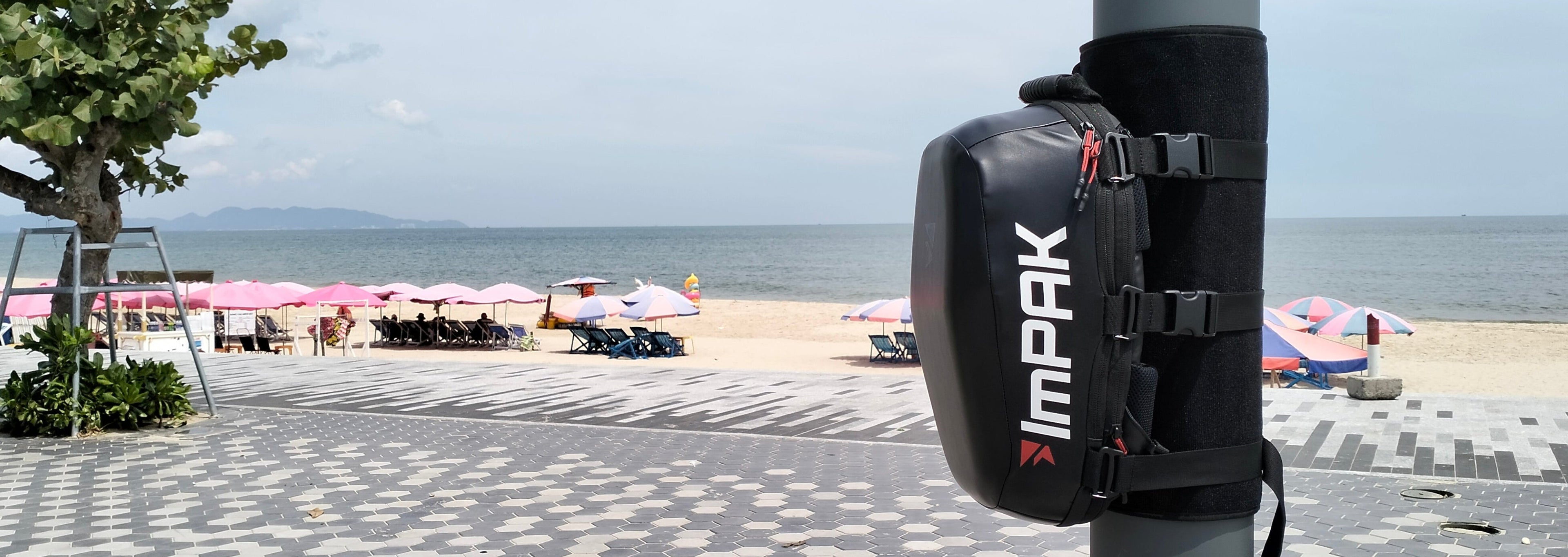 Pole with a backpack labeled 'imPAK' on a paved area near a beach.