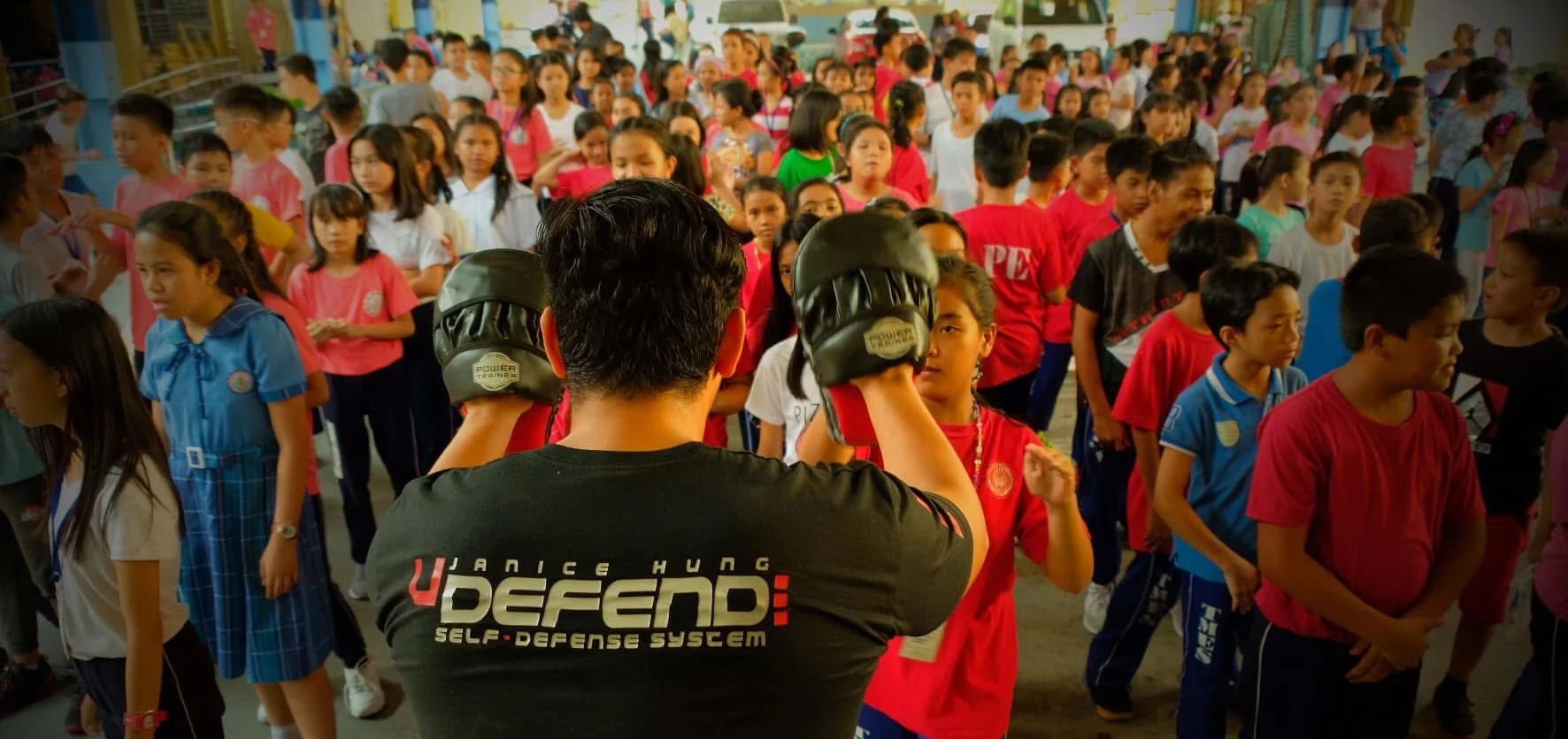 Group of people, mostly children, participating in a self-defense class with a instructor wearing a 'UDEFEND' shirt.